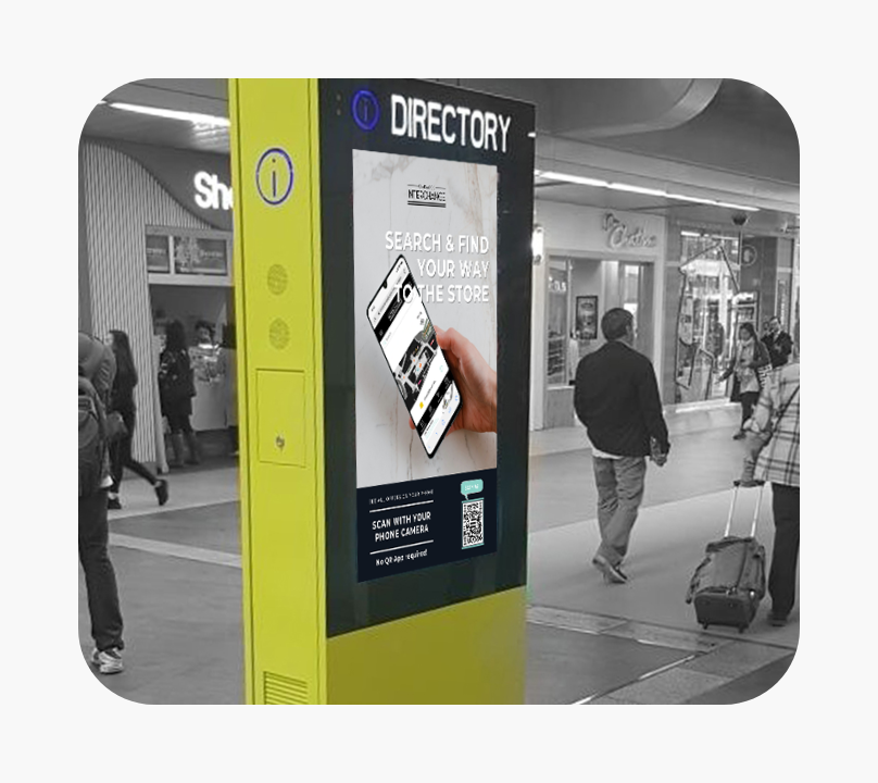 Yellow directory kiosk in a shopping mall displaying a screen with a hand holding a smartphone. Text on the screen reads 'SEARCH & FIND YOUR WAY TO THE STORE' and 'SCAN WITH YOUR PHONE CAMERA No QR app required' next to a QR code. People walk around in the background.