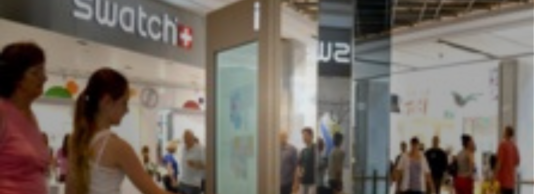 People walking and standing in a shopping mall corridor with a Swatch store visible in the background.