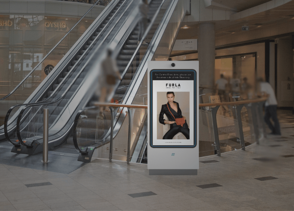 Digital advertisement kiosk in a shopping mall near escalators, showing a Furla ad featuring a woman in black attire holding a brown bag.