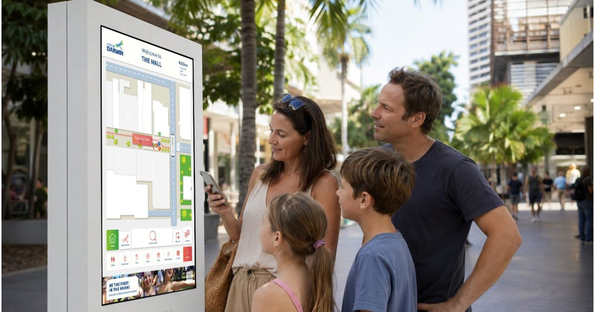 Family of four, including two children, looking at a digital city map kiosk outdoors with palm trees and city buildings in the background.