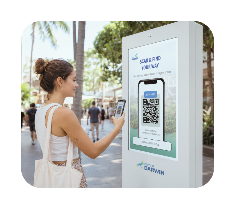 Woman with hair in a bun and a tote bag scans a QR code on a City of Darwin digital wayfinding kiosk outdoors on a sunny day with palm trees and people in the background.