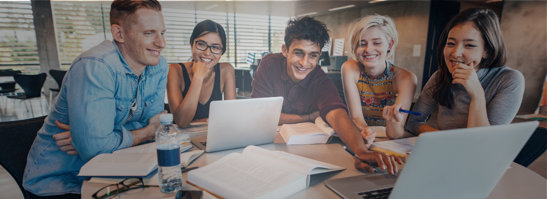 Five young adults sitting around a table, smiling and looking at two open laptops, with open books and a water bottle on the table, in a bright, modern room.