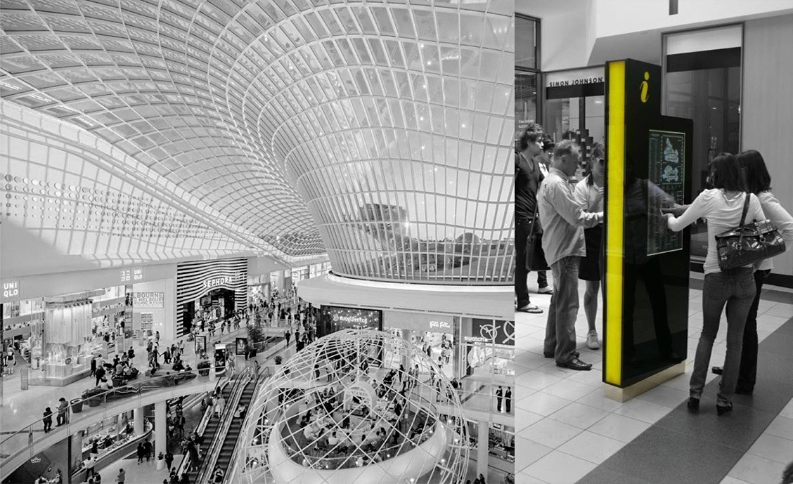 Inside a modern shopping mall with a large curved glass ceiling and multiple floors visible; an information kiosk with a yellow border and touchscreen is on the right where several people are interacting with it.