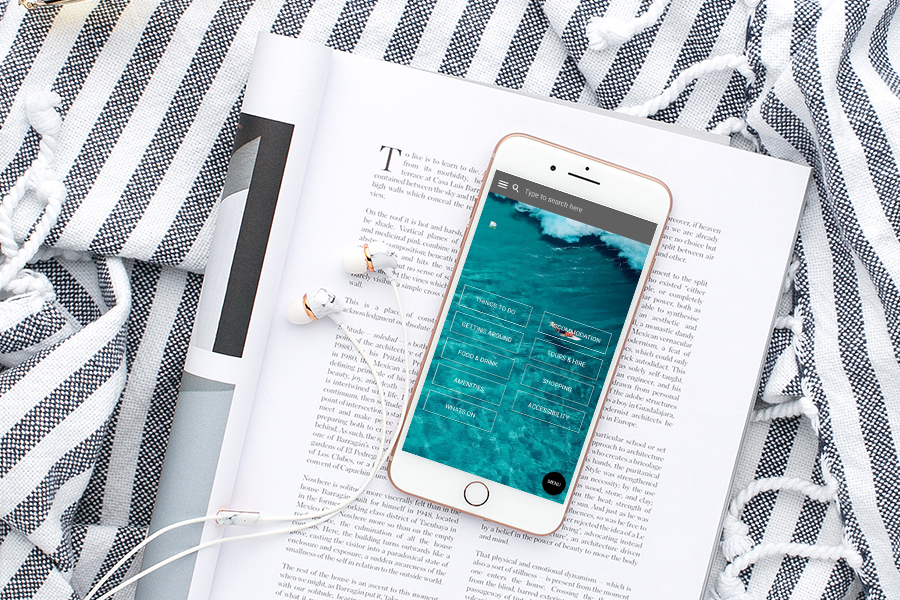 Smartphone with a clear ocean water background screen showing a menu with options like Things to Do, Accommodation, Getting Around, Tours & More, Food & Drink, Shopping, Amenities, Accessibility, and What's On, lying on an open book with visible text, placed on a white and black striped textured fabric with earphones plugged into the phone.
