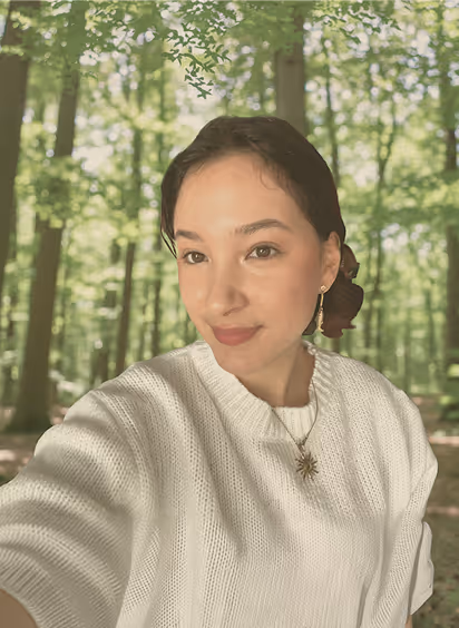 Woman wearing white sweater and sun-shaped necklace taking selfie in a green forest background.
