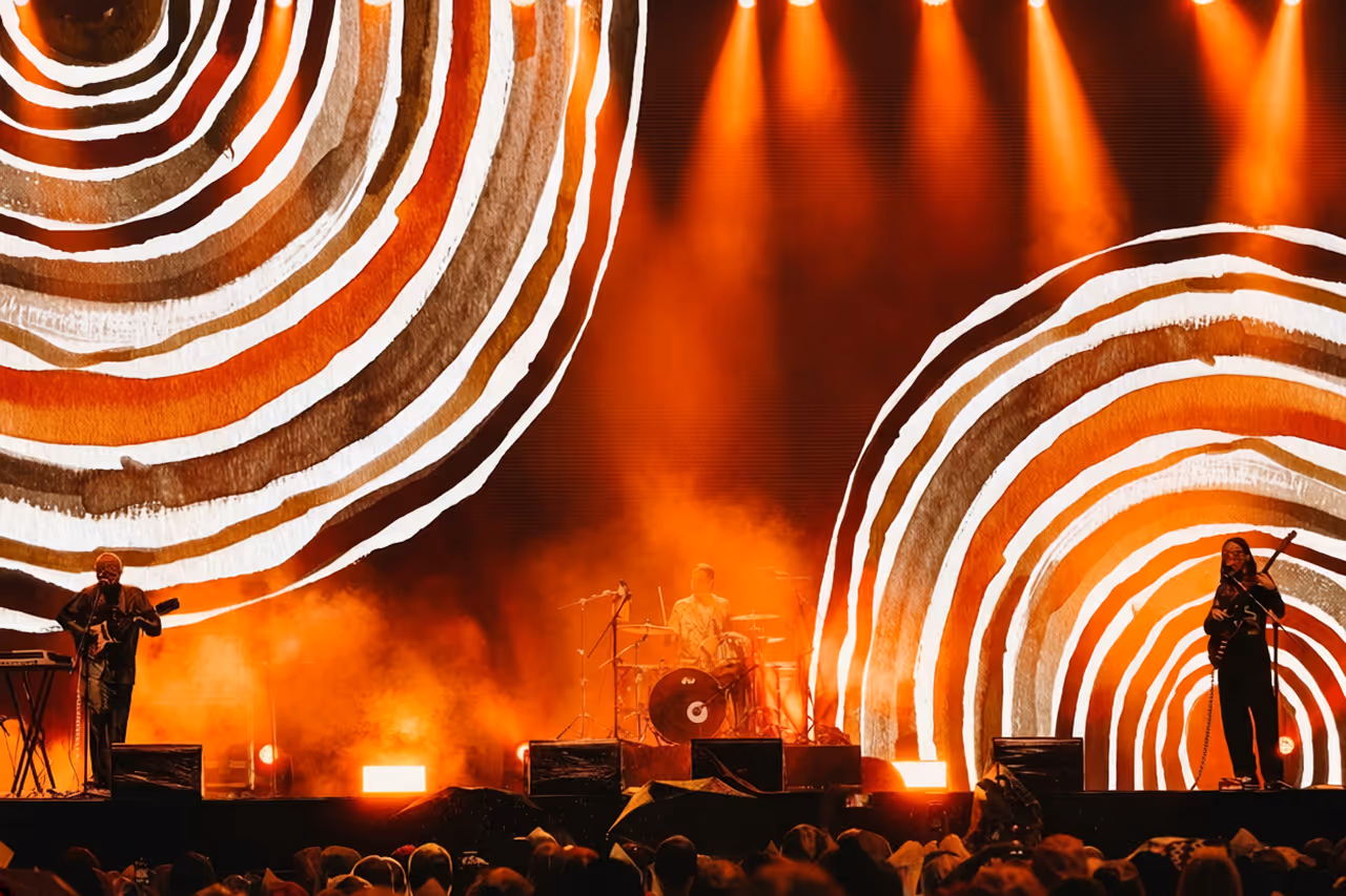 Band performing on stage with orange lighting and abstract circular artwork in background.
