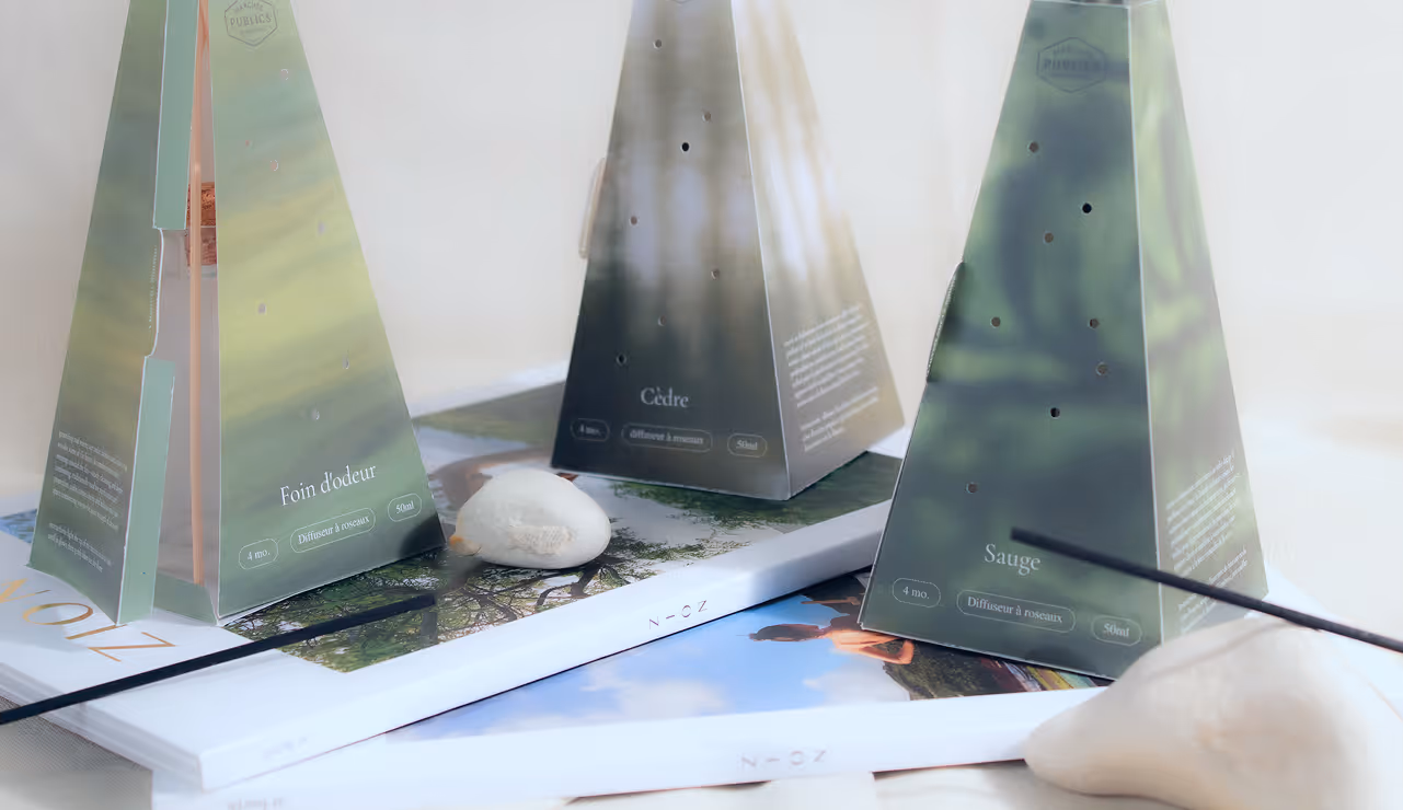 Three pyramid-shaped green diffuser boxes labeled Foin d'odeur, Cèdre, and Sauge on stacked magazines.