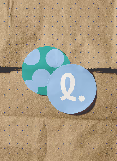 Two round stickers on a textured brown paper bag, one blue with a white stylized loop and dot, one green with blue circles.