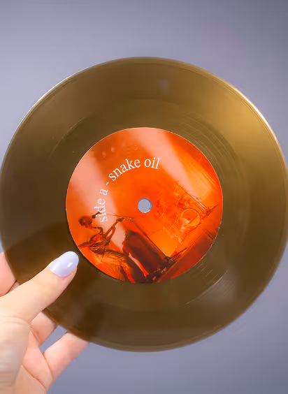 Hand holding gold vinyl record with orange label reading 'side a - snake oil'.