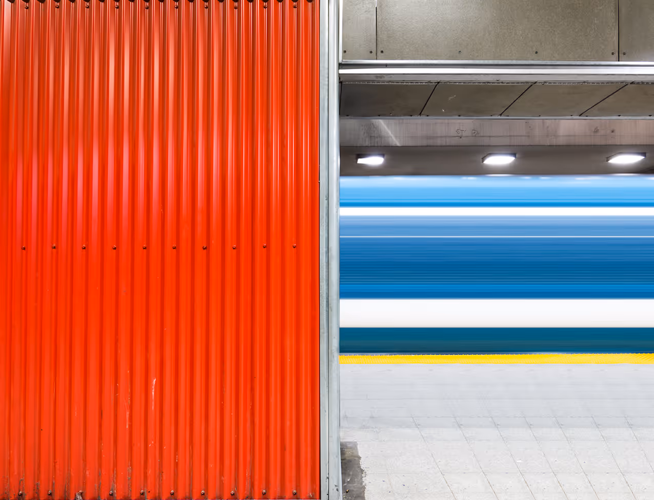 Blurred blue train passing a red corrugated metal wall at a subway station platform.