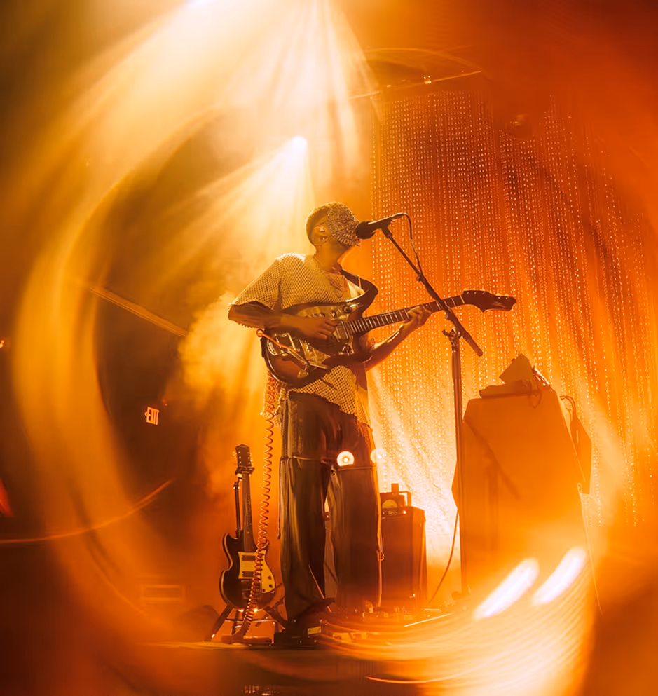 Musician wearing a beaded face mask playing electric guitar on stage bathed in warm orange light.