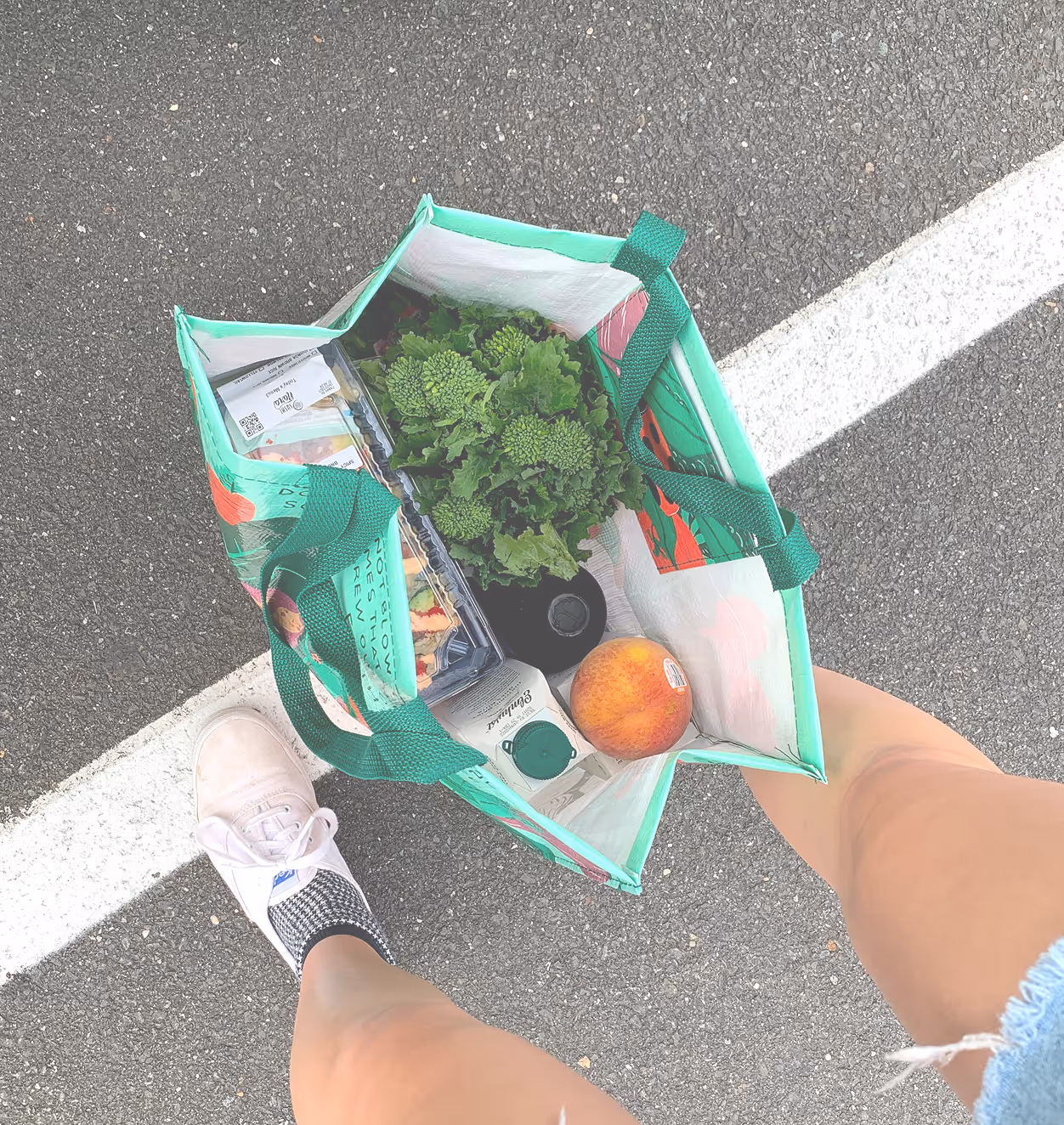 Person holding a grocery bag with lettuce, a carton, peach, and packaged food, standing on pavement.