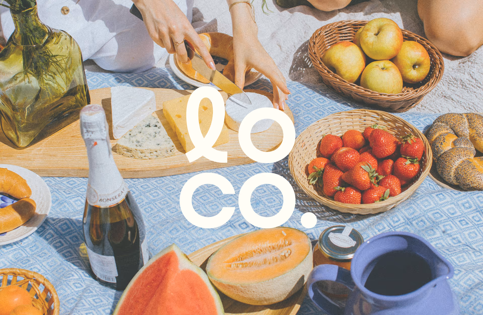 Picnic scene with cheese, fruits, bread, and a bottle on a blue cloth. On top, is the logo for the brand Loco.