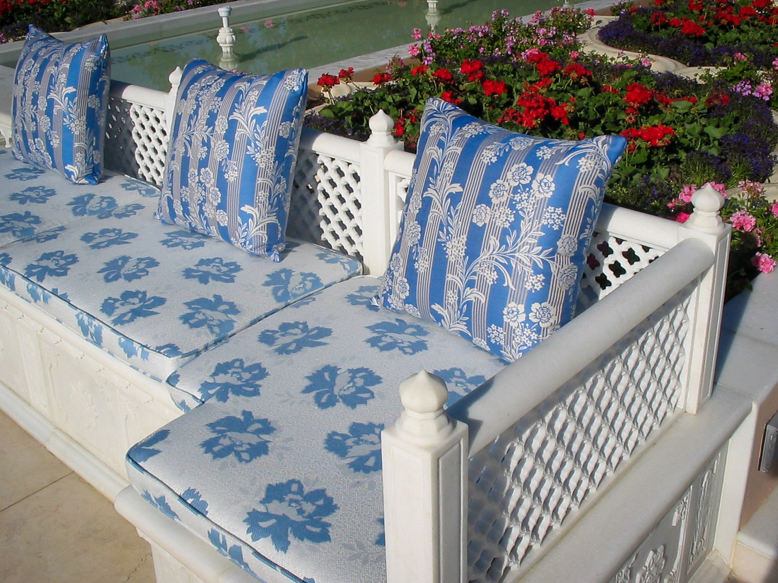 Handcrafted white marble garden bench featuring intricate jali lattice backrest and custom blue floral upholstered cushions, styled in a luxury estate outdoor setting
