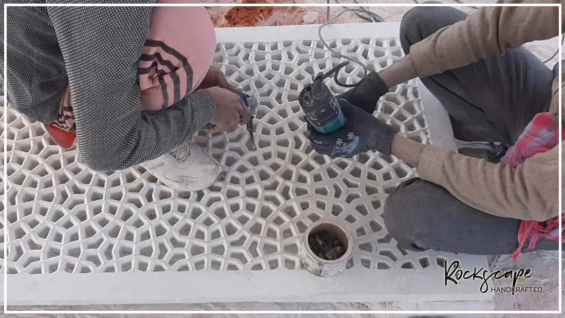 Two artisans hand-carving a large white marble jali panel in the workshop, seen from above