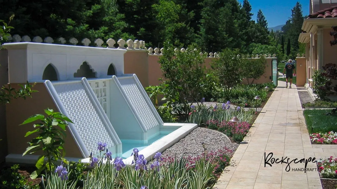 Marble water cascade flowing into a garden rill lined with flower beds and a stone path