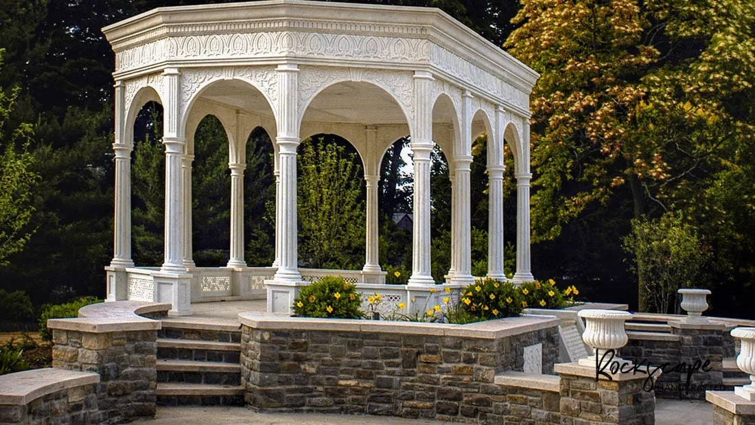Open white marble pavilion with arched columns on a stone terrace surrounded by trees