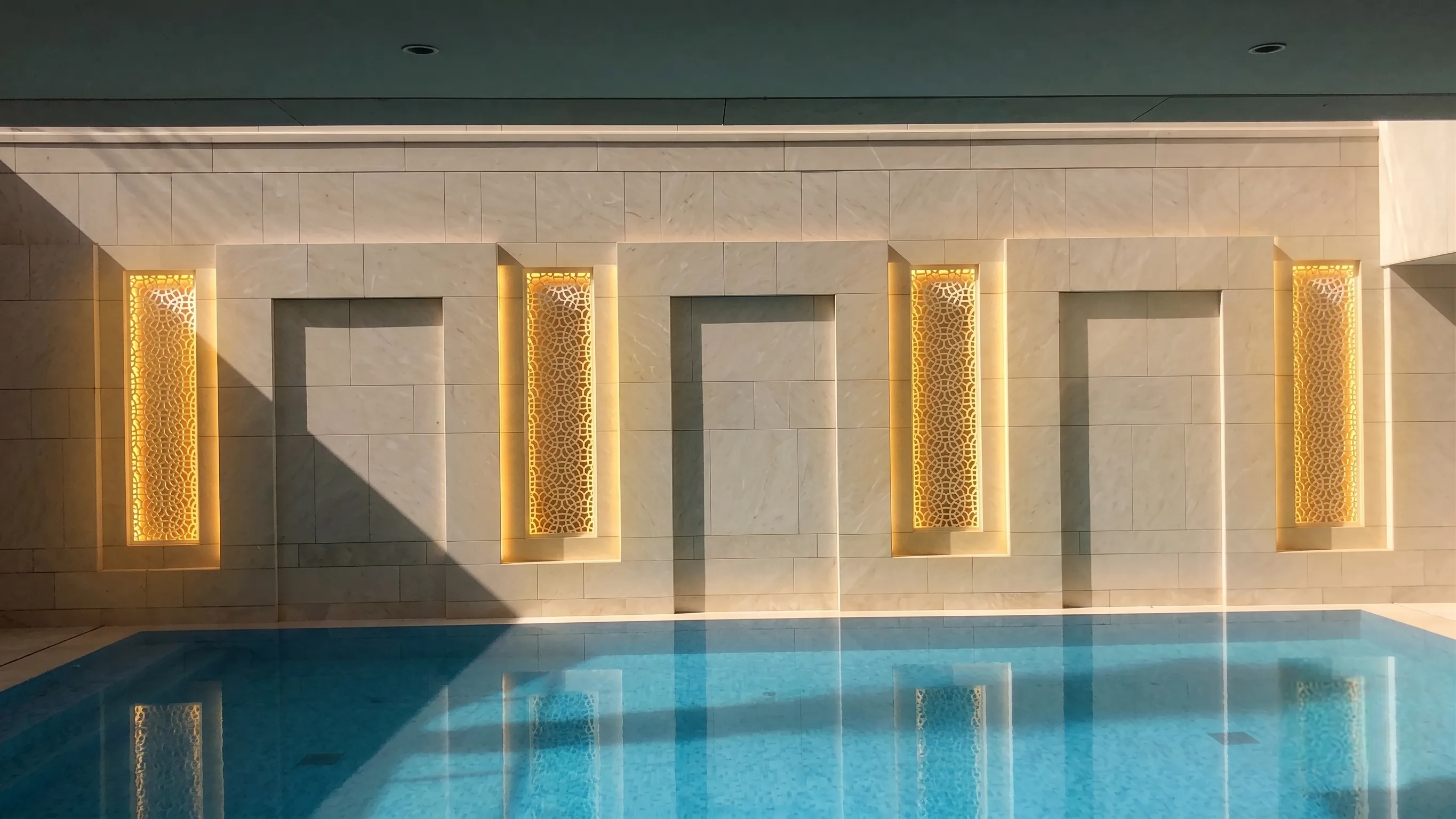 Indoor pool lined with a row of backlit marble jali niches along the stone wall, Al Mula Residence, Kuwait