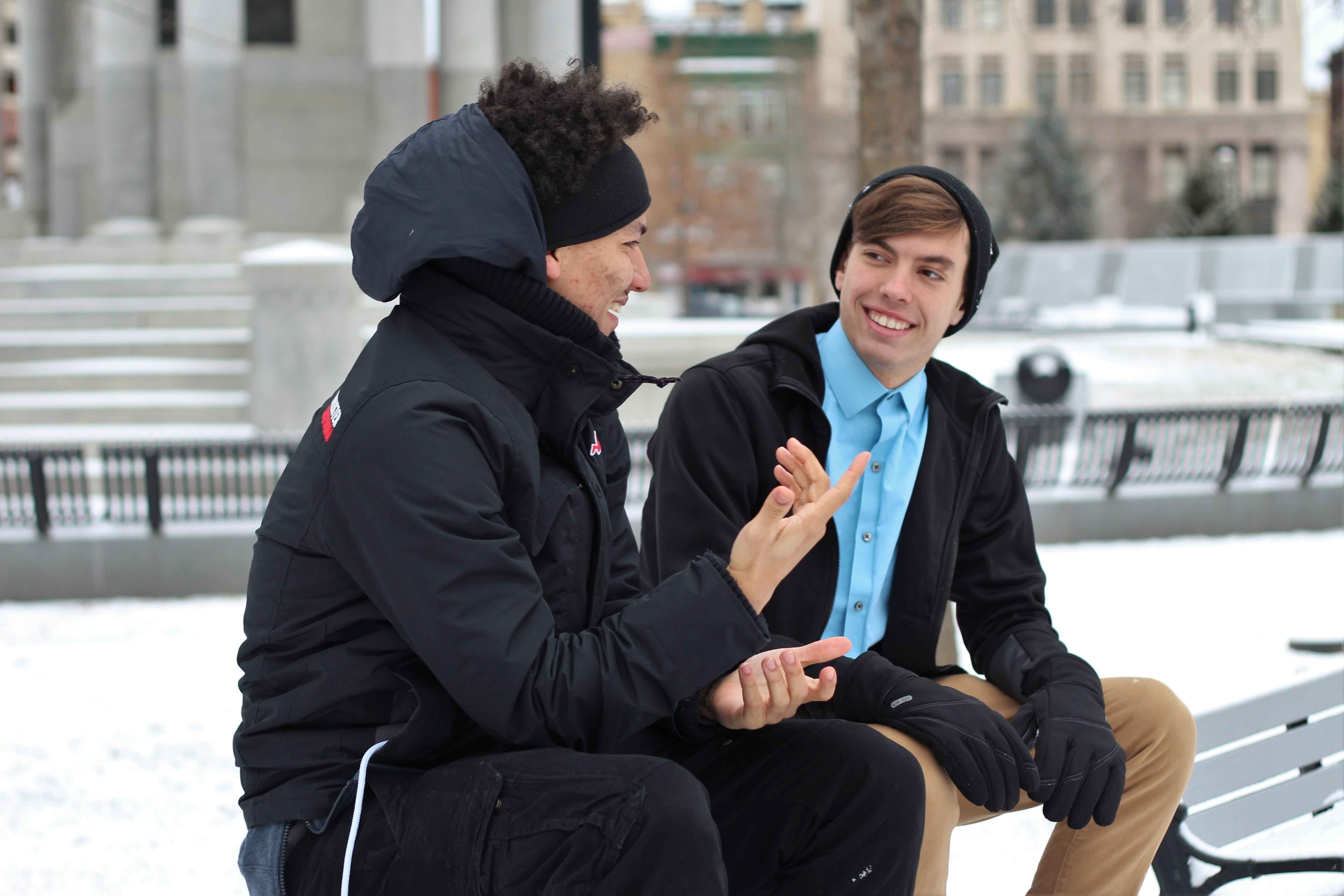Two men sitting on a bench talking in the snow|Man on his phone and woman smiling out the window while drinking coffee|Man giving a presentation to four colleagues|Woman shaking a mans hand with lady in background smiling