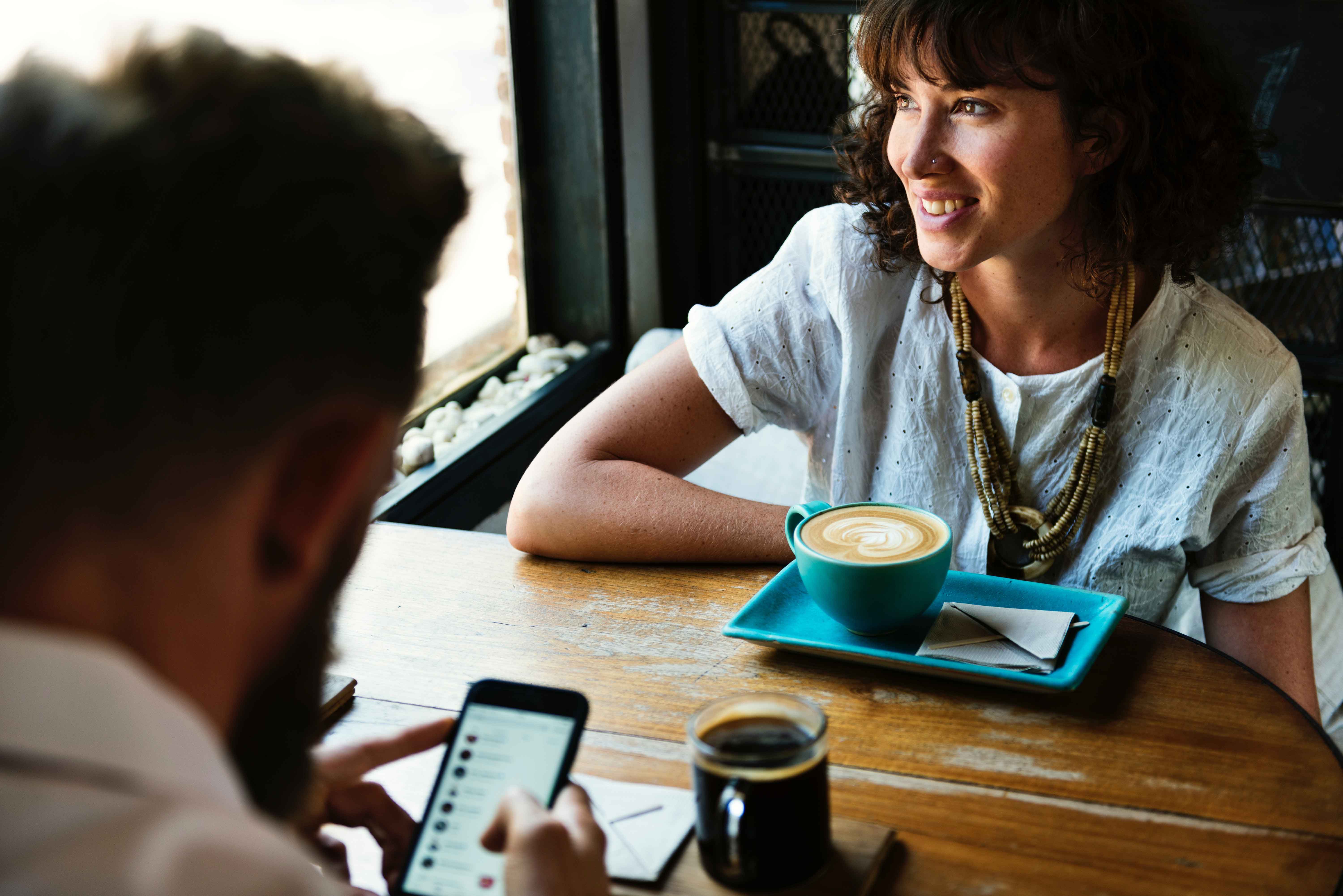 Man on his phone and woman smiling out the window while drinking coffee