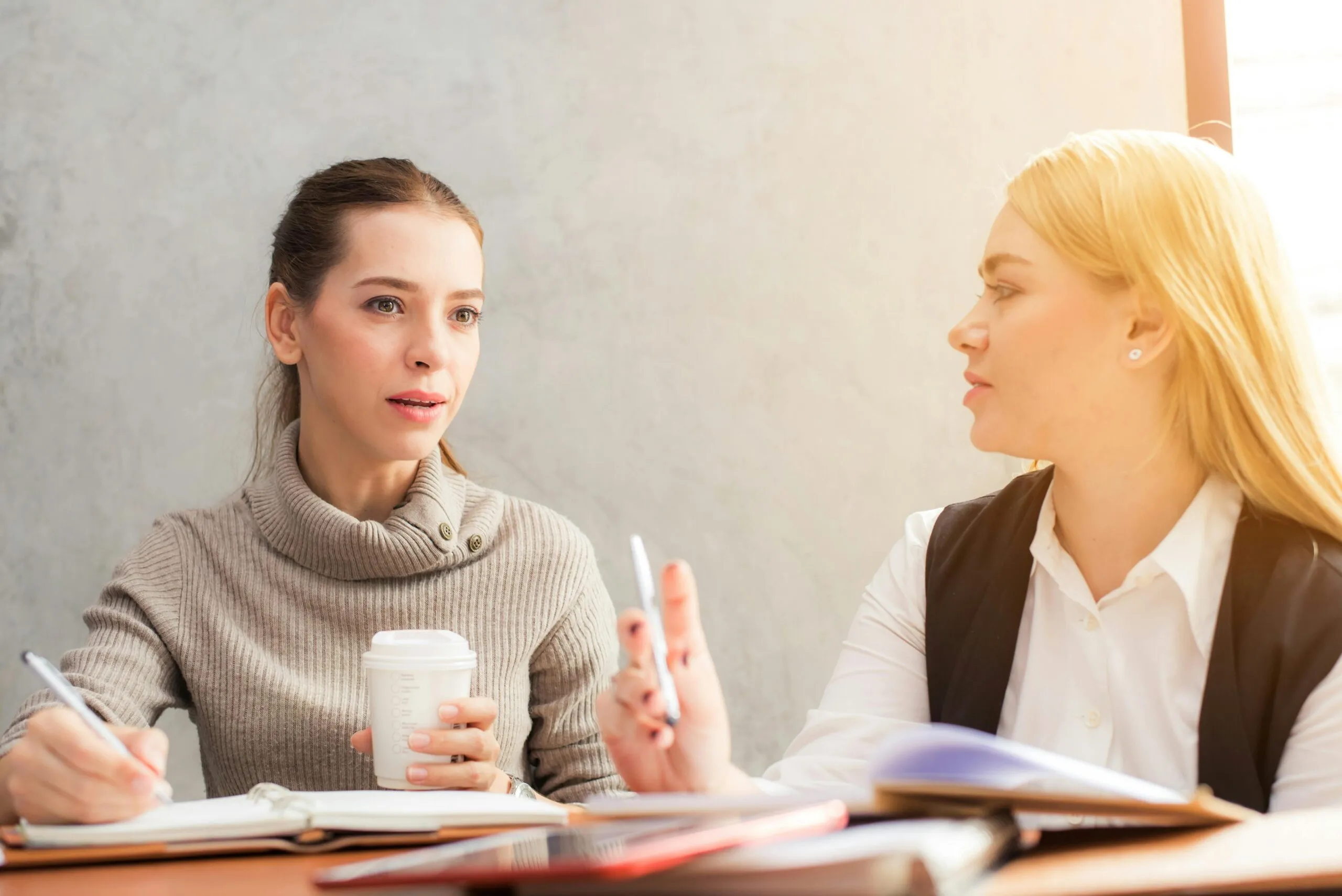 Two women are sitting at a table, engaged in conversation. The woman on the left, with dark hair in a beige turtleneck, holds a coffee cup and a pen with an open notebook in front of her. The woman on the right, with blonde hair in a white blouse and black vest, gestures with a pen as she speaks. They appear to be discussing something important.