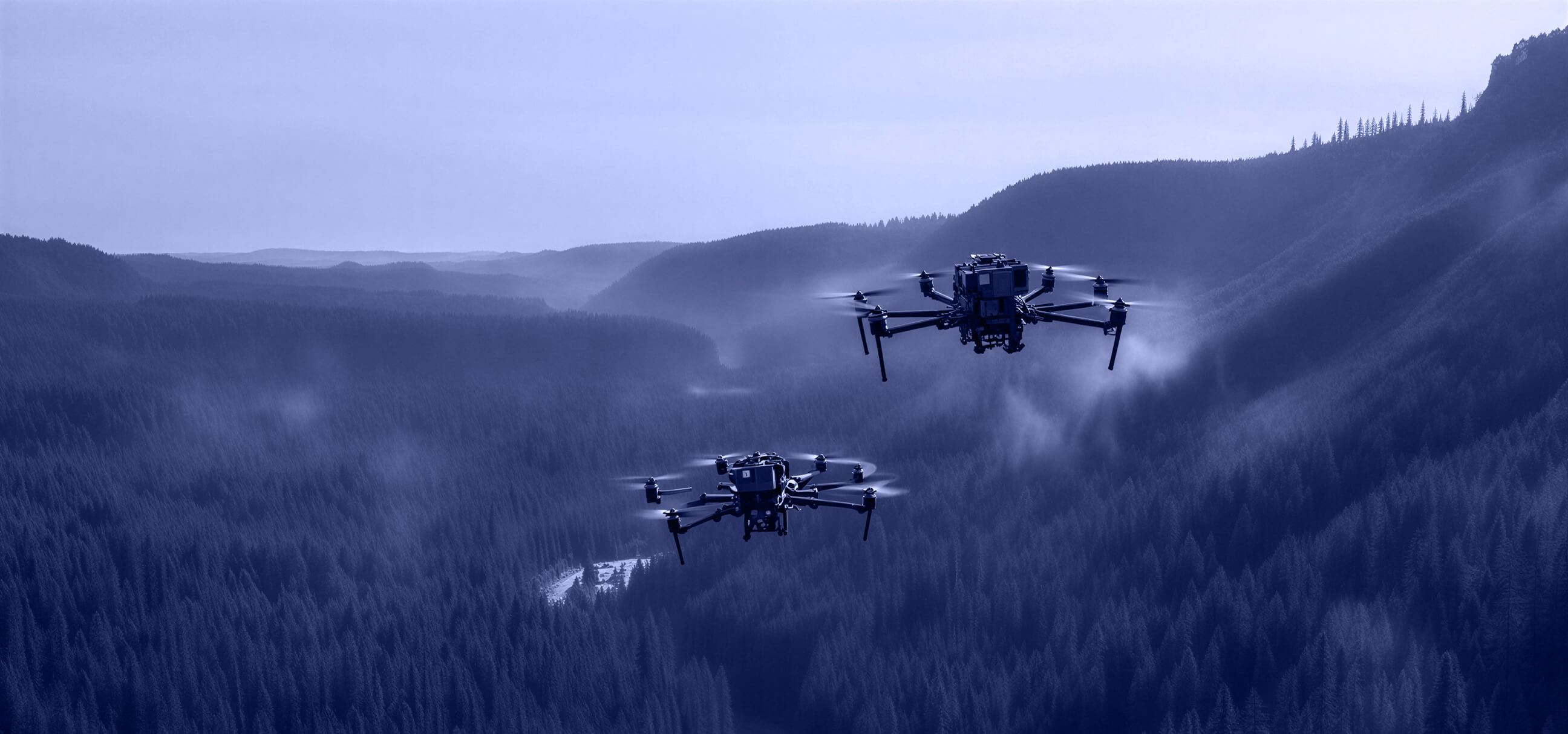 Two drones flying over a dense forest valley with mist and hills in the distance.