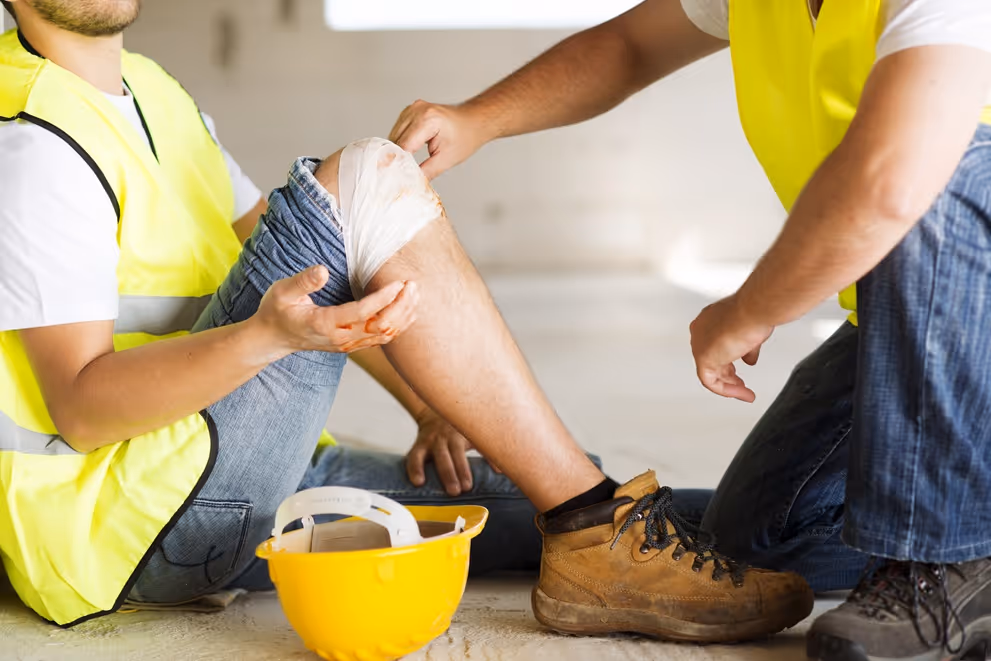 Worker in yellow vest helping another worker with a bandaged injured knee on a construction site.