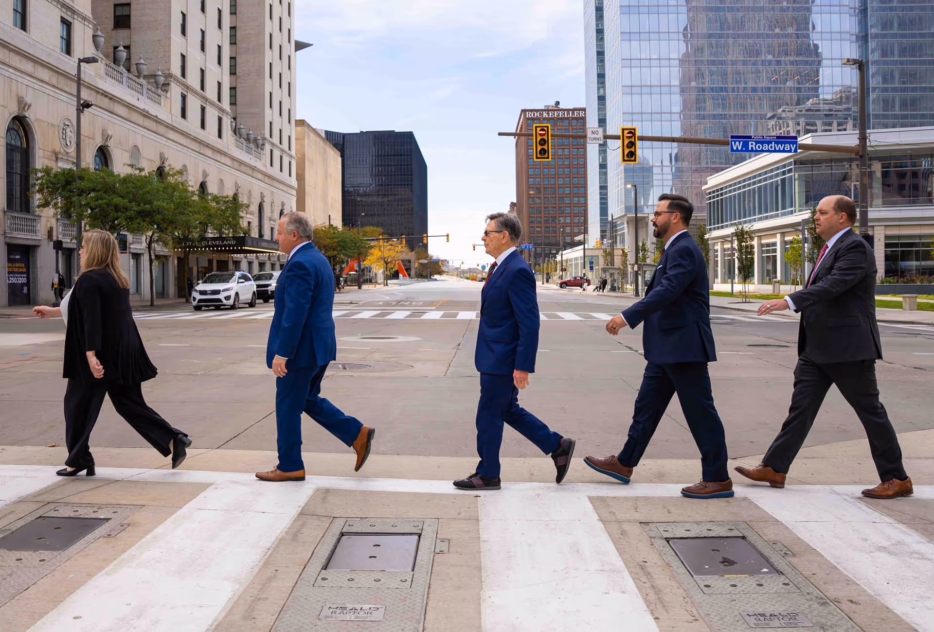 Five people walking across a city street crosswalk in business attire on a clear day.