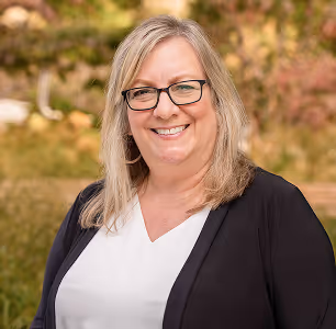 Smiling woman with glasses wearing a white top and black cardigan outdoors with fall foliage behind her.