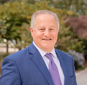 Smiling middle-aged man in a blue suit and purple tie standing outdoors with trees in the background.