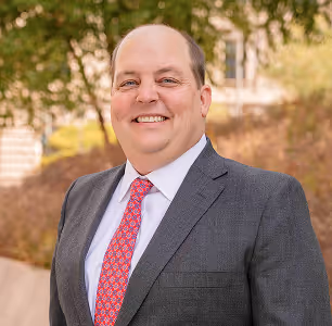 Smiling man in gray suit and red patterned tie standing outdoors with trees and buildings behind him.