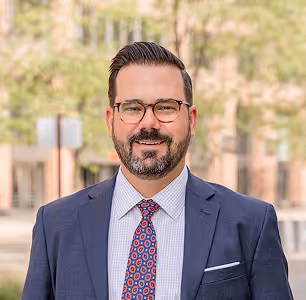Smiling man with glasses, beard, blue suit, and patterned tie standing outdoors.