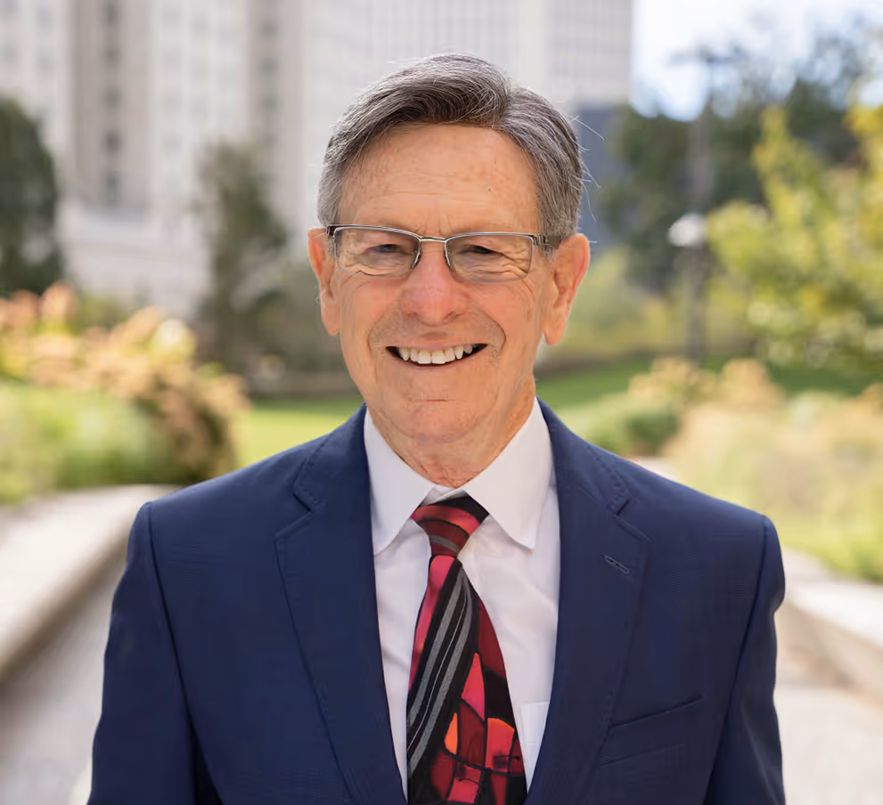 Smiling older man in glasses wearing a blue suit and colorful tie outdoors with blurred greenery behind.