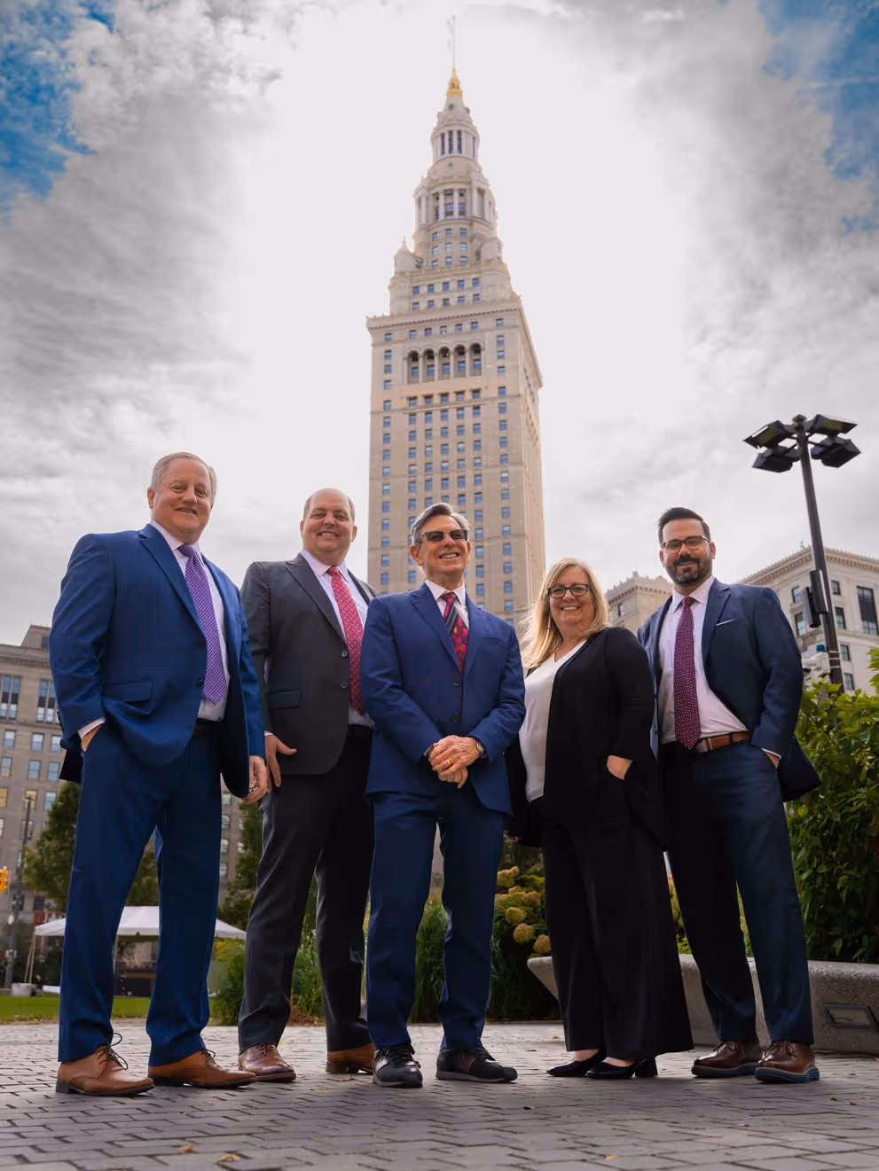 Group of five professionals in business attire standing outdoors with a tall building in the background.