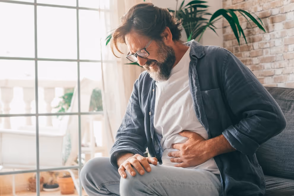 Man with glasses sitting on couch clutching stomach in pain near large window and plants.
