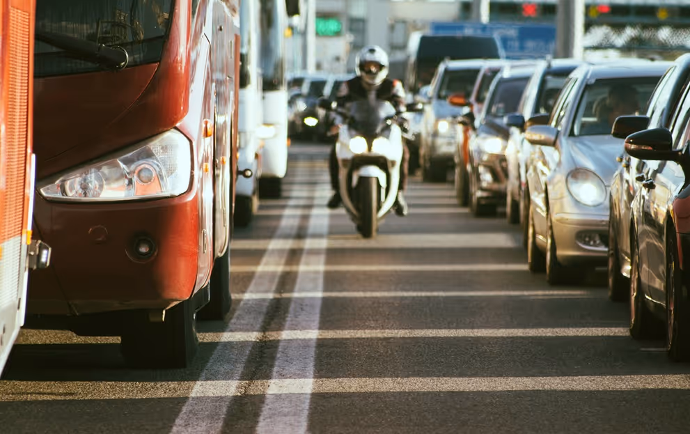 Motorcycle moving between lanes of stopped cars and buses on a busy road in daylight.