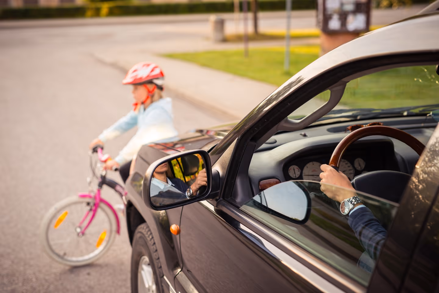 Child in red helmet riding pink bike near black car with driver holding steering wheel.