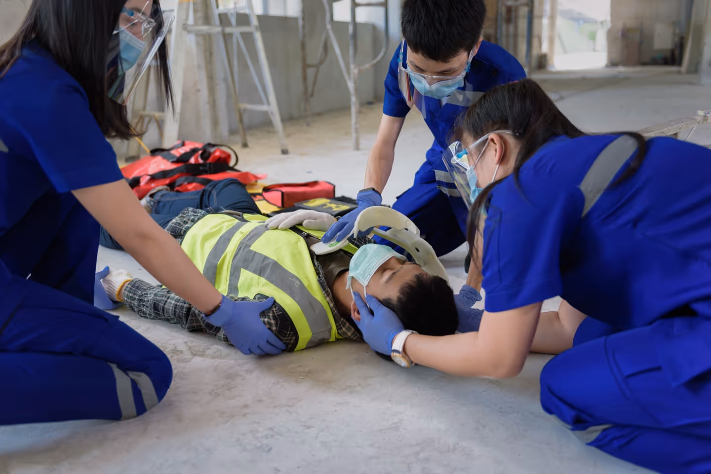 Three paramedics in blue uniforms attending to injured construction worker lying on floor with neck brace.