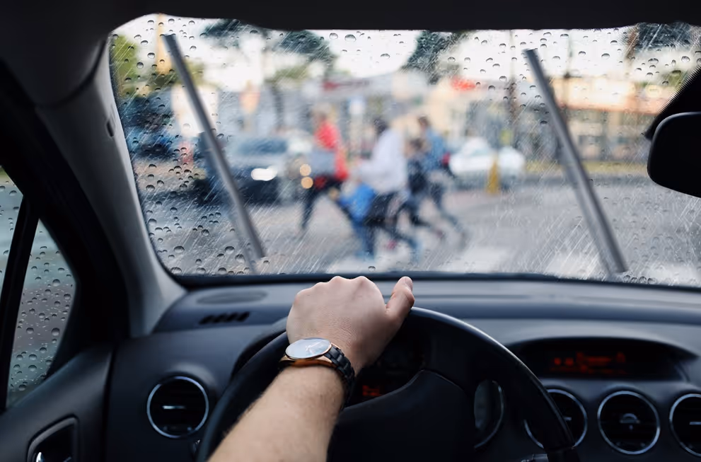 Driver's hand on steering wheel with wet windshield and pedestrians crossing street in background.