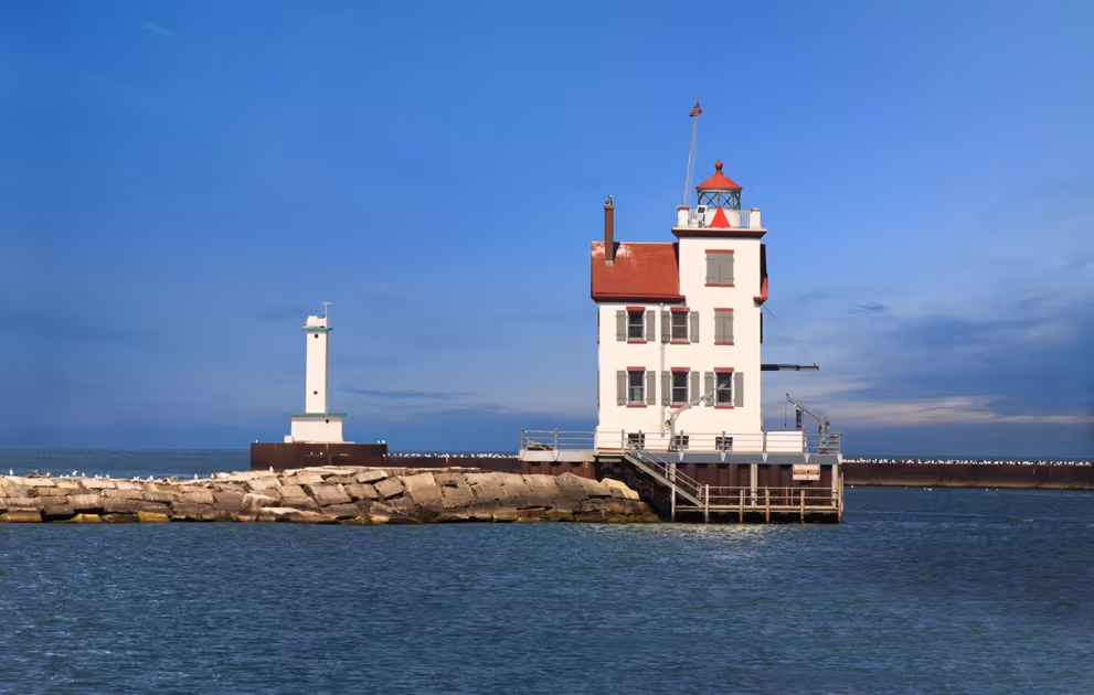 White lighthouse with red roof on a rocky pier extending into calm blue water under clear sky.