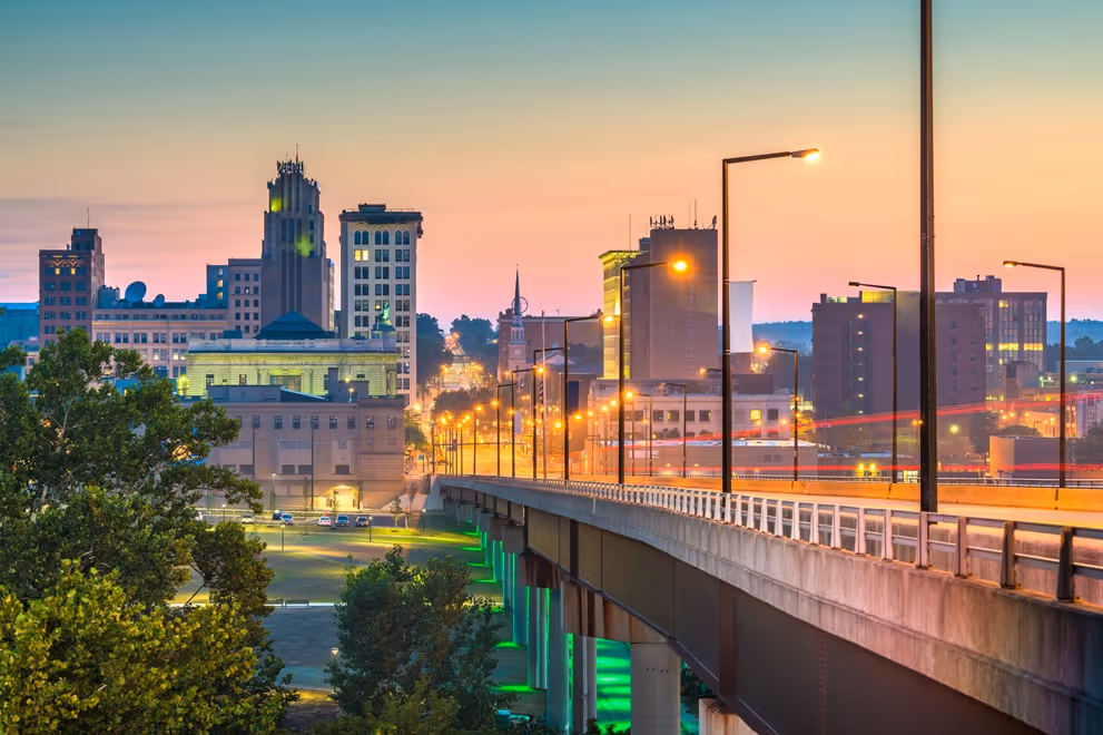 City skyline at sunset with lit streetlights and a bridge with green-lit supports in foreground.