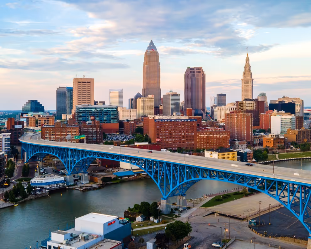Blue arched bridge over a river with Cleveland skyline and tall buildings at sunset.