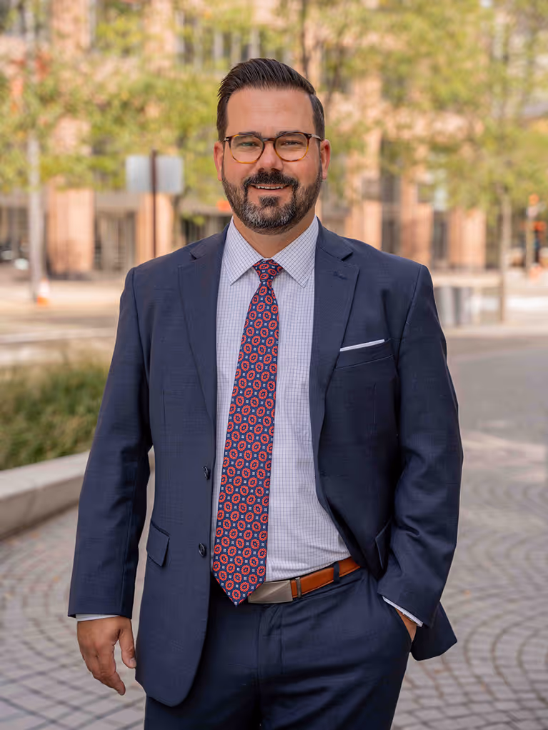 Smiling man with glasses wearing a dark blue suit and patterned red tie outdoors in an urban setting.