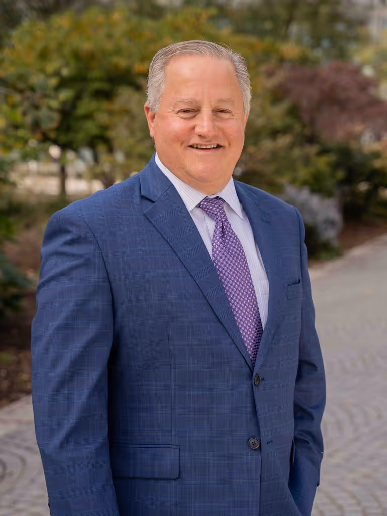 Smiling middle-aged man in a blue suit jacket and purple tie standing outdoors on a paved path.