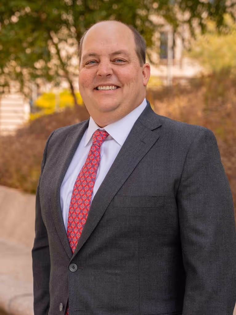 Smiling man in a gray suit and red patterned tie standing outdoors with trees in background.