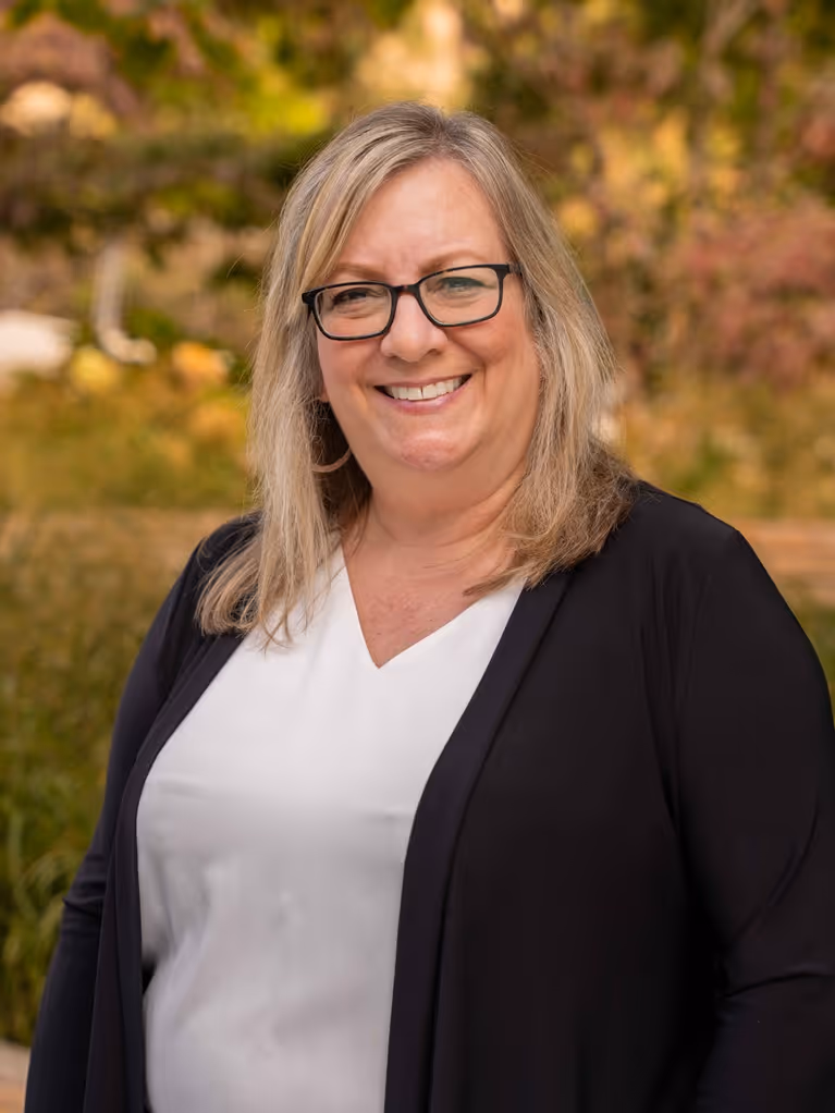 Smiling woman with glasses wearing black cardigan over white top outdoors with blurred foliage background.