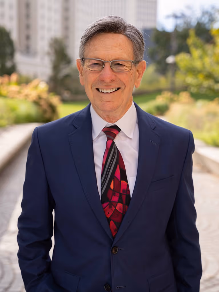 Smiling older man in glasses wearing a navy suit and colorful tie outdoors with blurred buildings and greenery.