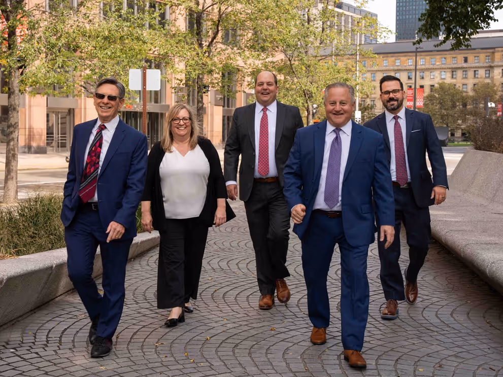 Five professionally dressed adults walking on a city sidewalk with trees and buildings in the background.
