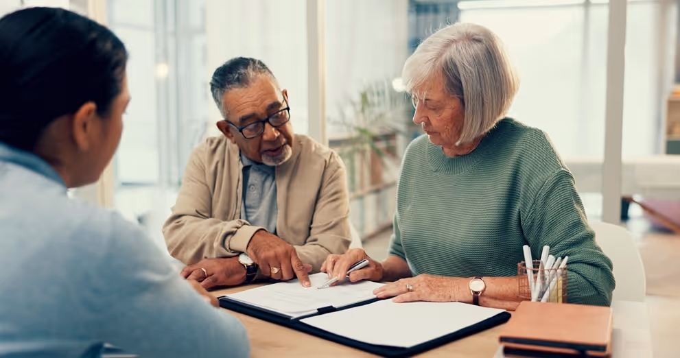 Senior man explaining documents to senior woman at desk with another person listening nearby.