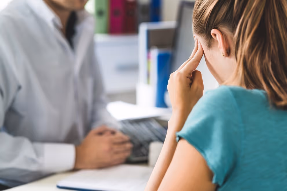 Woman sitting at a desk holding her head, talking to a doctor in a white coat.